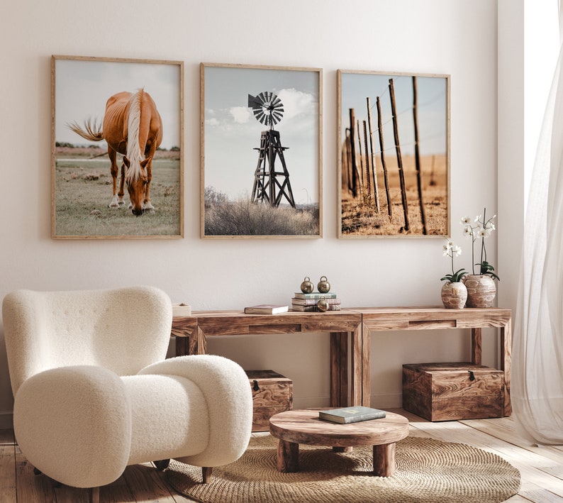 May include: Three framed prints on a white wall above a wooden table. The prints feature a brown horse grazing in a field, a windmill in a field, and a wooden fence in a field. The table has a wooden box on the left side and a wooden box on the right side. There is a small round wooden table in front of the table with a book on top.