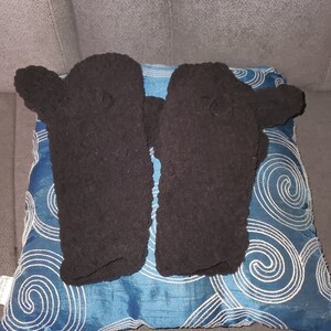 Puede incluir: Un par de guantes sin dedos de forro polar negro con un patrón de punto texturizado. Los guantes están colocados sobre una almohada azul y blanca con estampado.