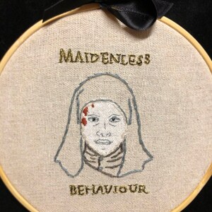 Puede incluir: Arte de bordado en aro con una figura blanca que lleva una cubierta para la cabeza y un cuello marrón. La figura tiene manchas rojas en la cara. El texto "MAIDENLESS BEHAVIOUR" está bordado en hilo dorado.