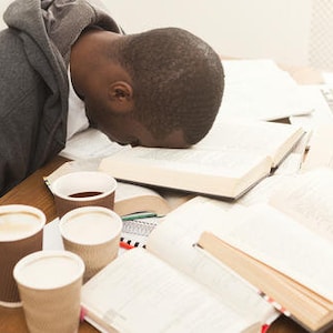 May include: A young person is sleeping face down on a table with several open books and a laptop. There are four disposable coffee cups on the table.