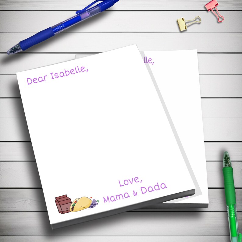 May include: A stack of white notepad paper with purple text that reads "Dear Isabelle," and "Love, Mama & Dada". The top sheet has the word "ille," written on it. There is a cartoon illustration of a milk carton, a taco, and grapes on the bottom sheet.