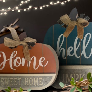 May include: Two wooden pumpkin-shaped signs with a rustic, farmhouse style. The orange sign says "Home Sweet Home" and the blue sign says "hello pumpkin".