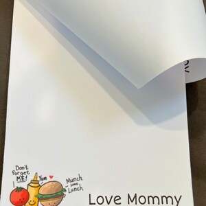 May include: White notepad with a cartoon illustration of a tomato, ketchup bottle, and hamburger with the text "Don't forget ME! Yum Munch some Lunch Love Mommy".