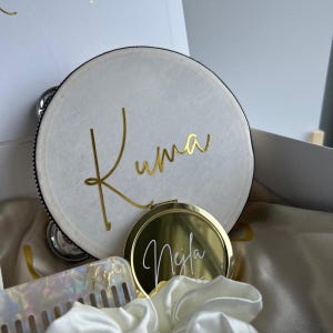 Könnte beinhalten: Ein weißes, kreisförmiges Etui mit dem Namen "Kuma" in goldener Schrift, ein goldener Kompaktspiegel mit dem Namen "Nejla" und ein perlmuttfarbener Kamm mit "Nejla" in goldener Schrift. Die Artikel sind Teil eines Geschenksets.