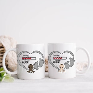 May include: Two white ceramic mugs with a gray heart design and the text "Loading... AUNT" with a progress bar filled with red hearts. One mug features a cartoon baby boy with brown skin and the other mug features a cartoon baby boy with white skin.