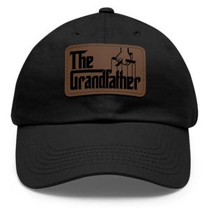 May include: Black baseball cap with a brown leather patch featuring the text "The Grandfather" and a puppet master graphic. The cap has a curved brim and a fabric strap closure.