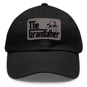 May include: Black baseball cap with a gray patch that says "The Grandfather" with a hand holding strings above the text.