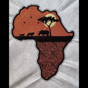 May include: An embroidered patch shaped like the continent of Africa. The patch is orange with a black outline and features a silhouette of a rhino, two lions, a tree, and a yellow sun. The bottom of the patch is a brown and black patterned design.
