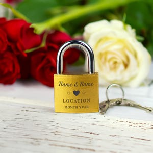 May include: A gold padlock with a silver shackle. The padlock is engraved with the words "Name & Name", two heart symbols, "LOCATION", and "MONTH YEAR". Keys are next to the padlock. Red and white roses are in the background.