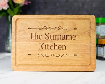 Puede incluir: Una tabla de cortar de madera con el texto "The Surname Kitchen" grabado en ella. La tabla tiene un acabado de madera natural y una forma rectangular.