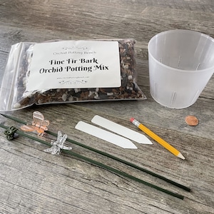 May include: A clear plastic pot, a bag of brown orchid potting mix, two white plant labels, a yellow pencil, and two green plant stakes with butterfly and dragonfly decorations. The bag of potting mix has a label that reads "Fine Fir Bark Orchid Potting Mix".