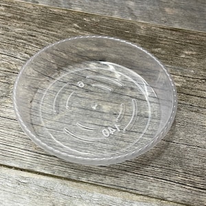May include: A clear plastic plant saucer with a 140 mm diameter. The saucer has a raised lip and a textured bottom.