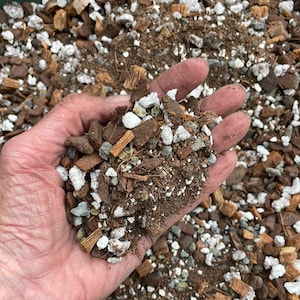 May include: A close-up of a hand holding a mixture of potting soil, coconut coir, and small white rocks. The soil is a dark brown color and the rocks are a light gray color.