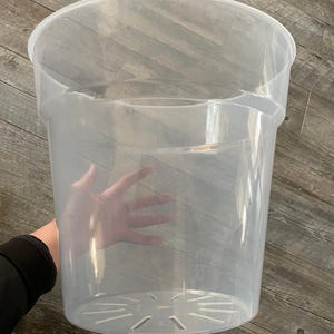 Pode incluir: Um vaso de plantas de plástico transparente e transparente, com uma borda larga e orifícios de drenagem na parte inferior. O vaso é cilíndrico com uma forma ligeiramente cónica. O vaso está vazio e pronto para plantar.