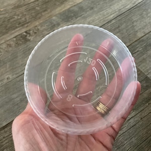 May include: Clear plastic saucer for a plant pot, with a diameter of 150 mm. The saucer has a raised rim and a drainage pattern on the bottom.