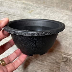 Könnte beinhalten: Ein schwarzer Plastik-Blumentopf mit einer strukturierten Oberfläche. Der Topf hat eine abgerundete Form und einen leicht ausgestellten Rand.