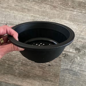 May include: A black plastic plant pot with a round shape and a perforated insert. The pot has a slightly textured surface and a wide rim. The pot is held by a hand with gold rings.