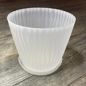 Könnte beinhalten: Ein weißer, durchscheinender Blumentopf aus Kunststoff mit passender Untertasse. Der Topf hat ein strukturiertes, wirbelndes Design und eine leicht ausgestellte Form. Der Topf ist für Pflanzen und Blumen konzipiert.