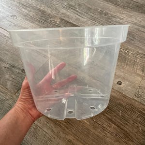 May include: A clear, transparent plastic plant pot featuring a wide, circular rim and a reinforced base with multiple drainage holes. The container has a smooth, tapered cylindrical shape designed for plant cultivation and root visibility.