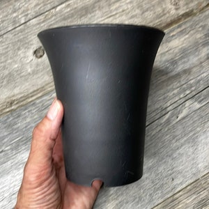 May include: A black plastic plant pot with a flared rim. The pot is smooth and has a slightly textured surface.