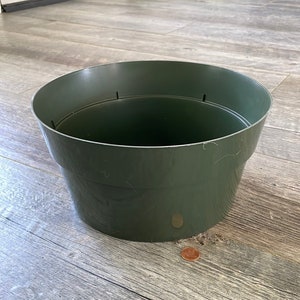 Puede incluir: Una maceta de plástico verde oscuro con una parte superior ancha y abierta. La maceta es redonda con una forma ligeramente cónica y tiene varios pequeños orificios de drenaje cerca del borde. Una moneda está al lado de la maceta para comparar el tamaño.