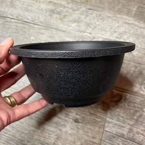 Puede incluir: Una maceta de plástico negra con una superficie texturizada y un borde ligeramente acampanado.
