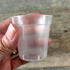 Puede incluir: Un vaso de plástico transparente y translúcido con un aspecto ligeramente esmerilado. El vaso es cilíndrico con un borde acampanado y pequeños pies en la base. El vaso se sostiene en una mano.