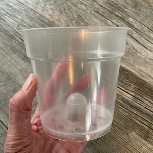 Peut inclure: Un pot de fleurs en plastique transparent et incolore, doté d'un cône central arrondi surélevé à la base et de multiples trous de drainage autour du périmètre du fond. Le récipient présente un bord évasé et une structure cylindrique lisse.