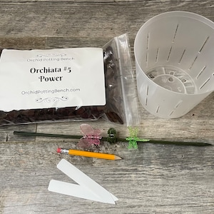 May include: A set of orchid potting supplies including a bag of orchid bark, a white plastic pot with drainage holes, a green stake with a butterfly decoration, a pencil, and two white plant labels. The bag of orchid bark has a label that reads "Orchid Potting Bench, Orchiata #5 Power, OrchidPottingBench.com".