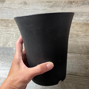 Op de afbeelding: Een zwarte plastic plantenpot met een gegolfde rand. De pot wordt in een hand gehouden.