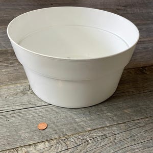 Puede incluir: Una maceta de plástico blanco con una abertura ancha y redonda y un cuerpo ligeramente cónico. La maceta está sobre una superficie de madera envejecida, con una moneda visible para la escala.