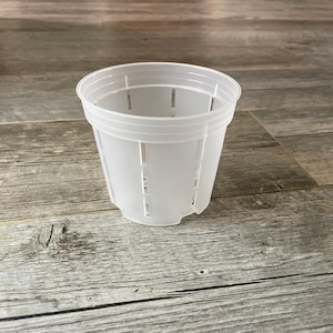 May include: A clear, plastic plant pot with a cylindrical shape. The pot has vertical slits for drainage and ventilation. The pot is designed for planting flowers, herbs, or other small plants. The pot is sitting on a wooden surface.