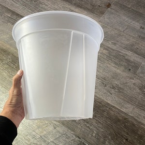 May include: A translucent white plastic planter with a wide, rounded rim and a faceted design. The planter is held by a hand, showcasing its size and shape. The container is designed for plants.