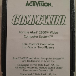 May include: A black Atari 2600 Commando video game cartridge. The label features the Activision logo and the game title in green. Text on the label includes game details and copyright information.
