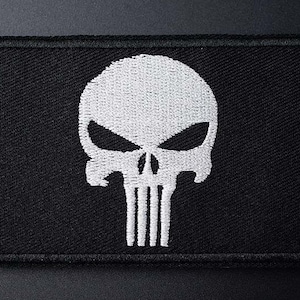 May include: Black rectangular patch featuring a white skull design. The skull has large eye sockets and vertical lines for teeth. The patch is made of a textured fabric.