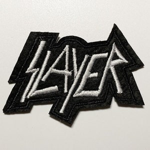 May include: Black embroidered patch with the word "SLAYER" in white, stylized lettering. The patch has a unique, angular shape and is likely designed to be sewn onto clothing or accessories. The patch is on a white surface.