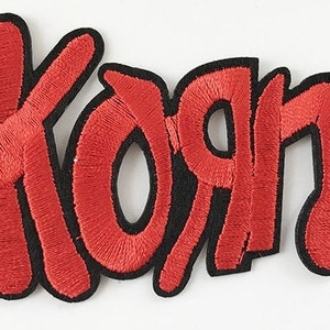 May include: Embroidered patch featuring the word "Korn" in red, stylized lettering. The letters are outlined in black, creating a bold contrast. The patch is likely designed to be sewn or ironed onto clothing or accessories.