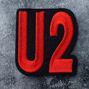 May include: Embroidered patch featuring the letters "U2" in bright red against a black background. The patch has a square shape with rounded corners and is likely designed to be ironed or sewn onto fabric.