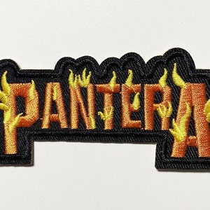 May include: Embroidered patch featuring the word "PANTERA" in orange letters with yellow flames. The patch has a black border with a scalloped edge. The design is likely for a band or music-related merchandise.