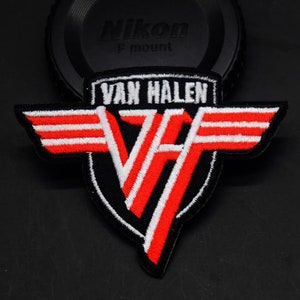 May include: Embroidered patch featuring the band name "Van Halen" in white lettering above a stylized "VH" logo. The logo is red and white, with black outlining and wings. The patch is likely for clothing or accessories.
