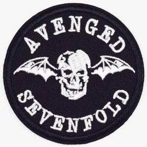 May include: A circular black patch with white embroidered text and a skull with bat wings. The text reads "AVENGED SEVENFOLD" in a curved font around the skull graphic. The patch has a stitched border.