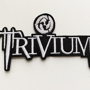 May include: A black and white embroidered patch featuring the band name "TRIVIUM" in white lettering. The patch includes a circular emblem above the band name. The patch is likely designed to be ironed or sewn onto clothing or accessories.