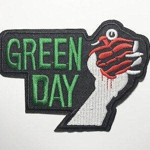 May include: Embroidered patch featuring the words "GREEN DAY" in green on a black background. The design includes a white hand clutching a red, claw-like object with blood dripping down.