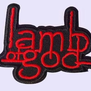 May include: Embroidered patch with the words "Lamb of God" in red script on a black background. The patch has a black border and is likely designed to be sewn or ironed onto clothing or accessories.