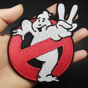 May include: Embroidered patch featuring the Ghostbusters logo. The design includes a white ghost character making a peace sign, set against a red circle with a diagonal red line. The patch has a black border.