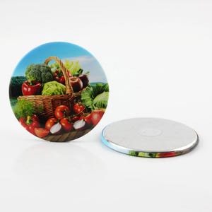 Puede incluir: Un imán de nevera redondo con una imagen colorida de una cesta de productos frescos, incluyendo tomates, brócoli y manzanas. El imán tiene un respaldo plateado.