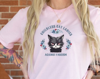 Op de afbeelding: Roze T-shirt met een zwarte kat afbeelding en de tekst "Childless Cat Ladies Against Fascism" in blauw.
