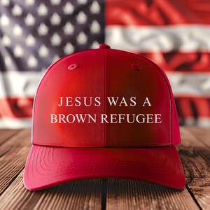 May include: A red baseball cap with the text "JESUS WAS A BROWN REFUGEE" in white. The cap is set against a backdrop of the American flag and a wooden surface.
