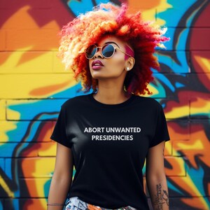 May include: A person with bright red, orange, and yellow hair is wearing a black t-shirt with the text "ABORT UNWANTED PRESIDENCIES" in white letters. The person is standing in front of a colorful graffiti wall.