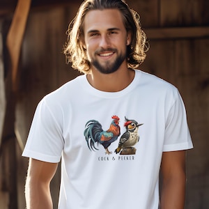 Puede incluir: Camiseta blanca con una ilustración de un gallo y un pájaro carpintero. El texto "Cock & Pecker" está impreso debajo de las aves.